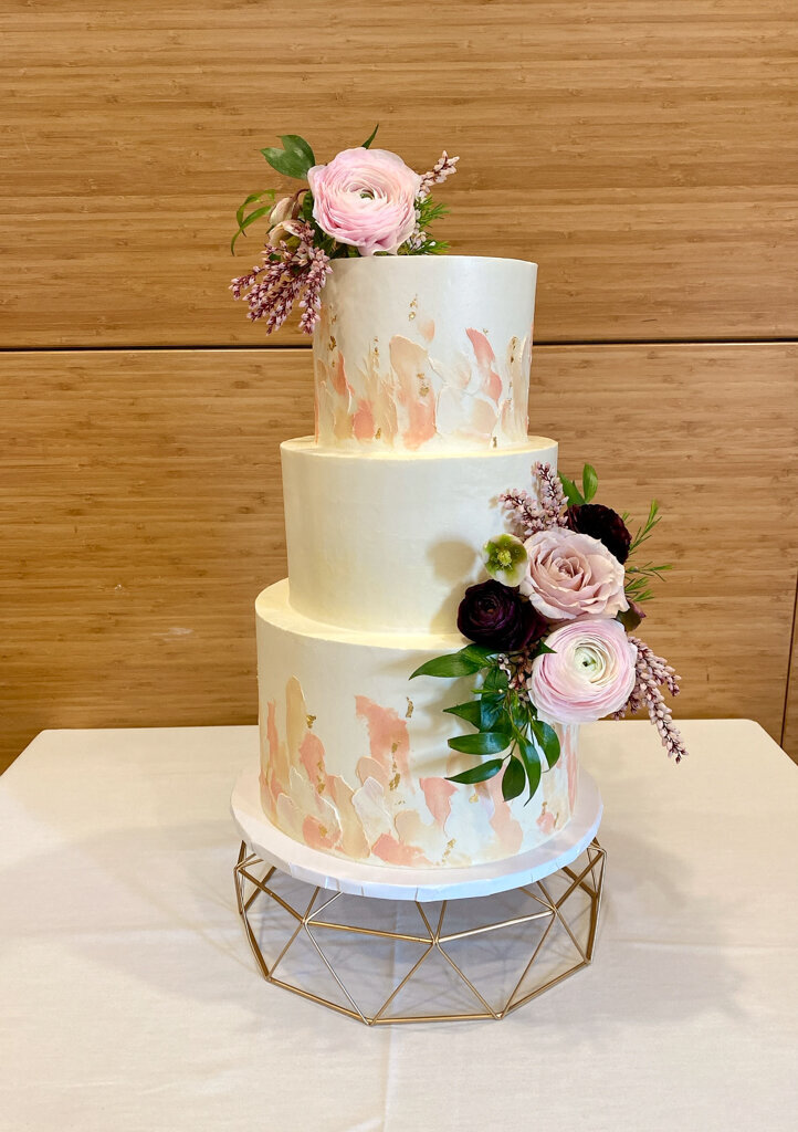 White 3-tiered wedding cake with pink and gold details and burgundy flowers, created by Caroline Lem, chef and owner of Lemonberry Pastries in Calgary, Alberta