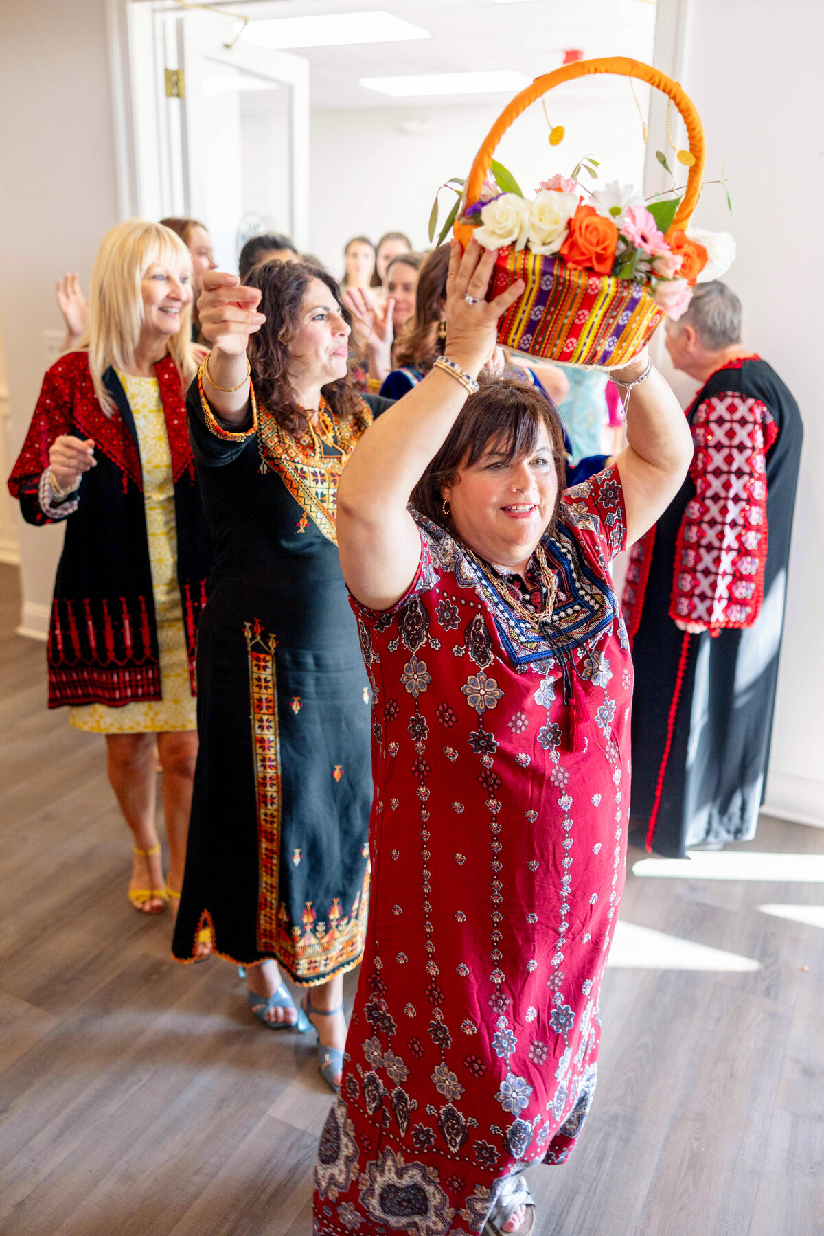 A woman in traditional attire carries a colorful basket of flowers on her head while leading a procession of similarly dressed women. They appear to be participating in a cultural or celebratory event indoors, with natural light illuminating the scene.