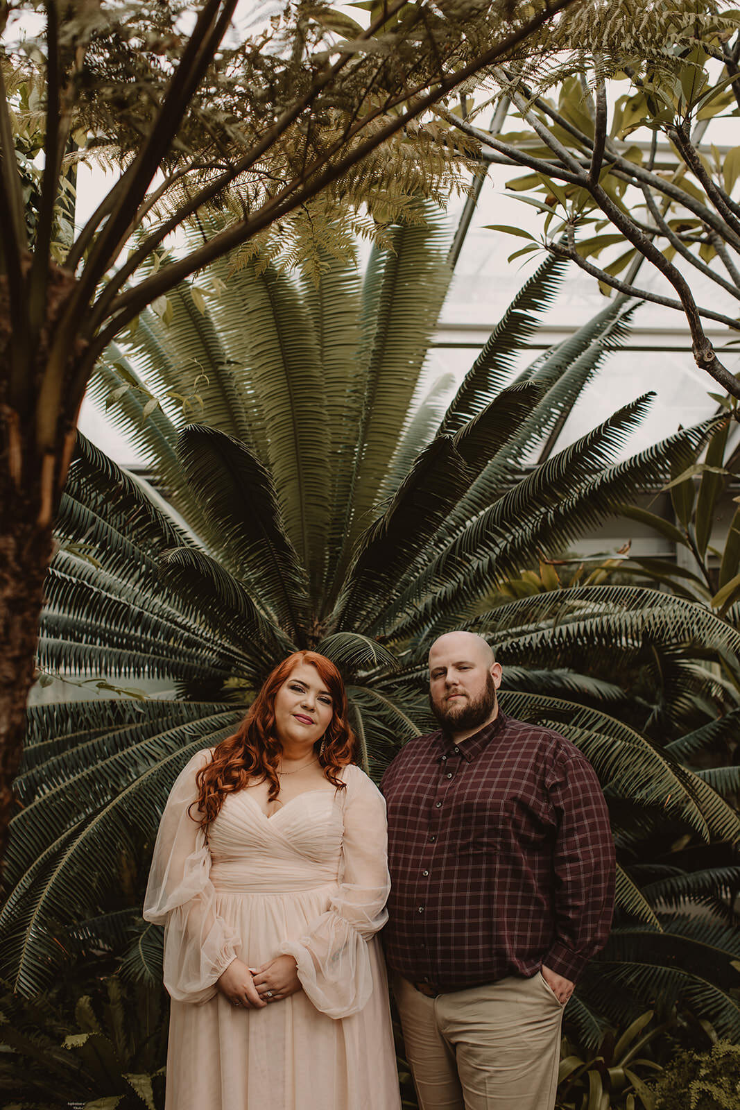 A couple standing shoulder to shoulder in front of plants in a conservatory 