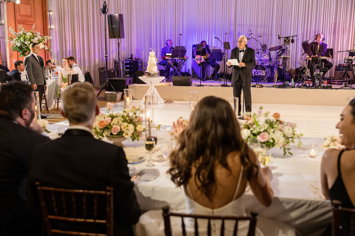 A man in a tuxedo is giving a speech at a wedding reception.
