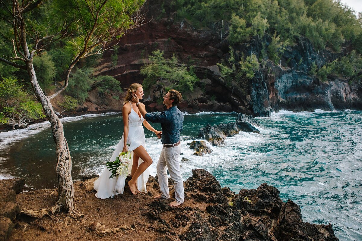 A bride and groom stand together on a cliffside overlooking the ocean during their Hawaii adventure elopement