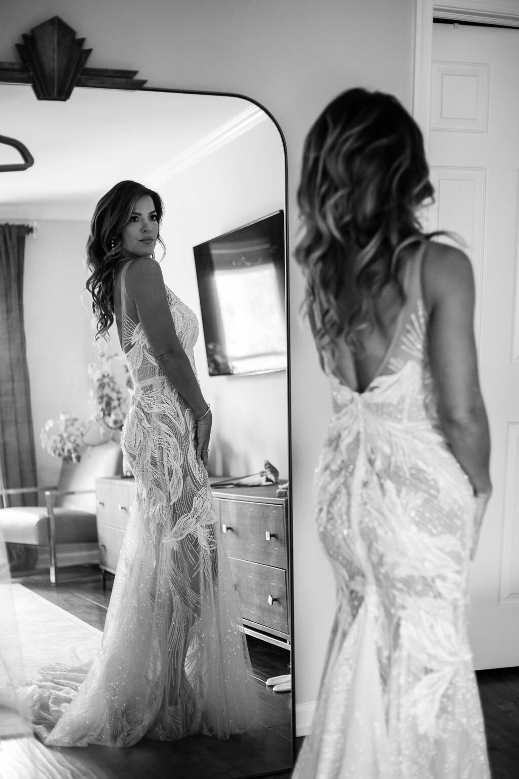 A mirror reflection of the bride as she admires her intricate lace wedding gown, standing gracefully in a warmly lit room.