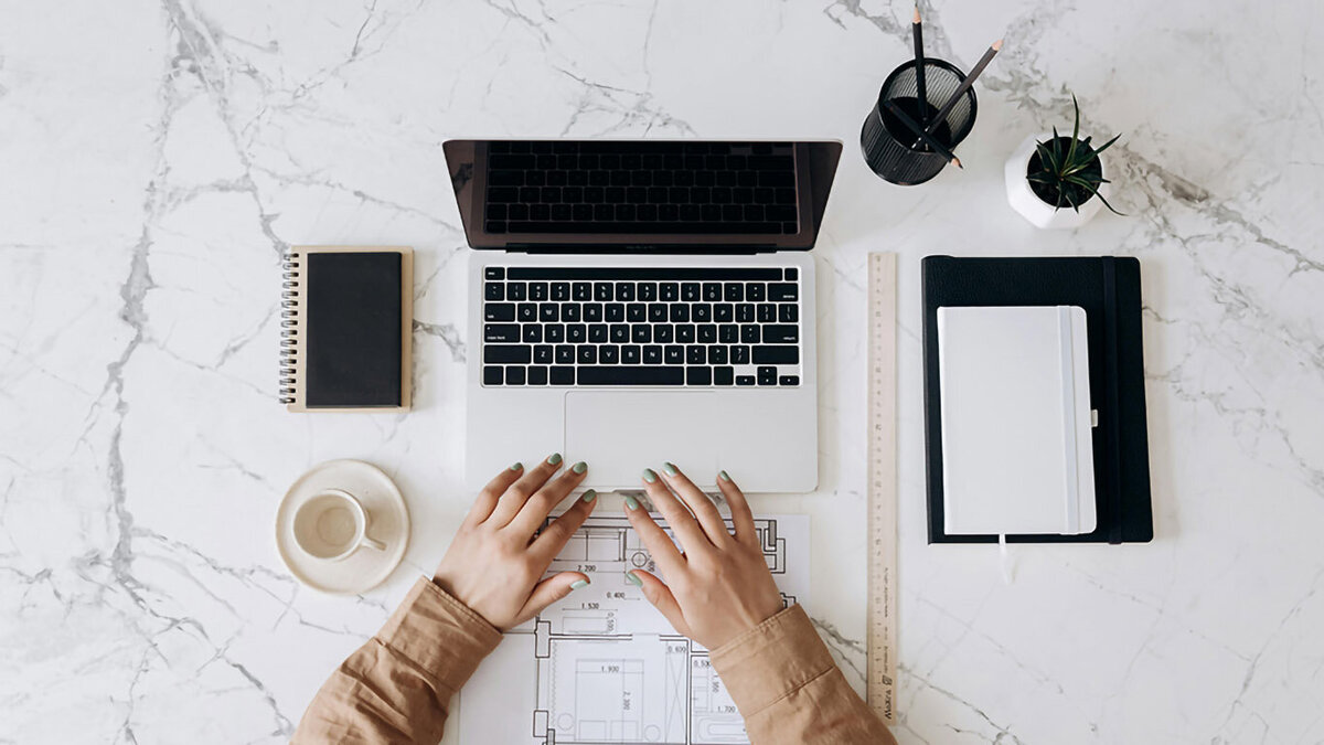 Overhead view of a workspace with hands on a laptop, coffee cup, notebook, and office supplies on a marble desk, symbolizing strategy and creative business support.