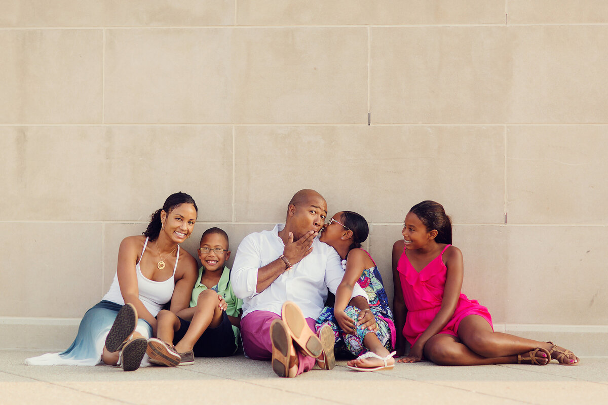 This family is sitting against a concrete wall in bright clothing. Dad is receiving a kiss from his daughter and he has a surprised, excited look on his face.
