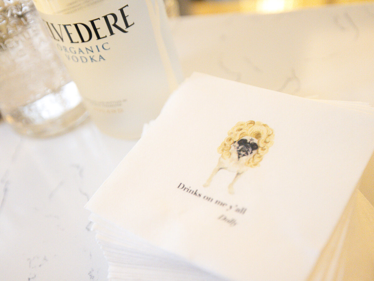 A stack of custom napkins sits on a marble surface next to a bottle of Belvedere Vodka. The napkin features an illustration of a pug in a wig, with the text Drinks on me yall, Dolly.