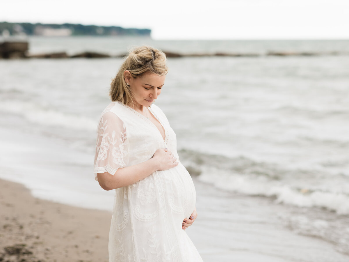 pregnant mother in white maternity gown holding baby bump on beach for maternity photoshoot cleveland maternity photographer