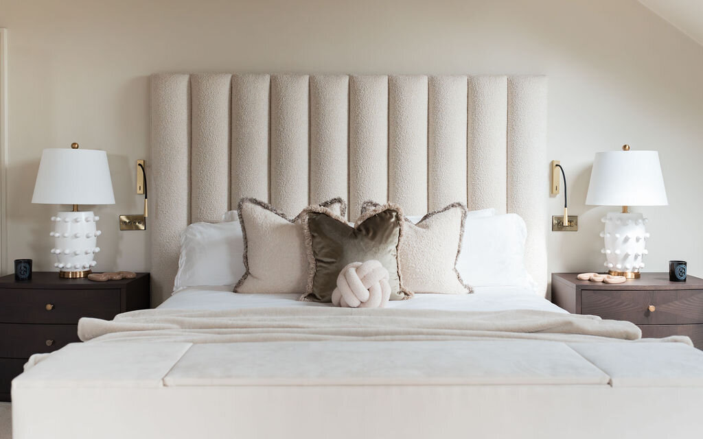 A well-lit bedroom featuring a large plush bed with a tall, tufted headboard. The bed is flanked by two dark wooden nightstands, each with a white lamp. The room has a neutral color palette with white and beige bedding and some accent pillows.