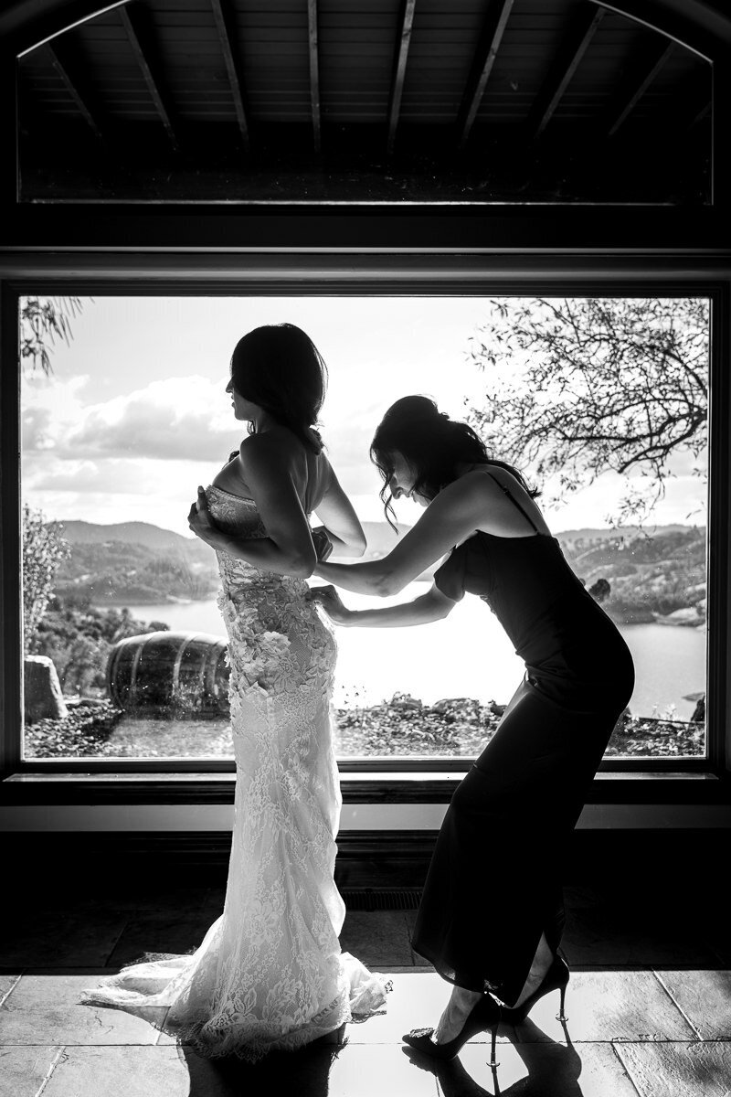 A heartfelt moment as the bride’s sister helps her button up her dress at Airola Road Vineyard, with a breathtaking view of the Sierra Nevada foothills framed through the large window behind them.
