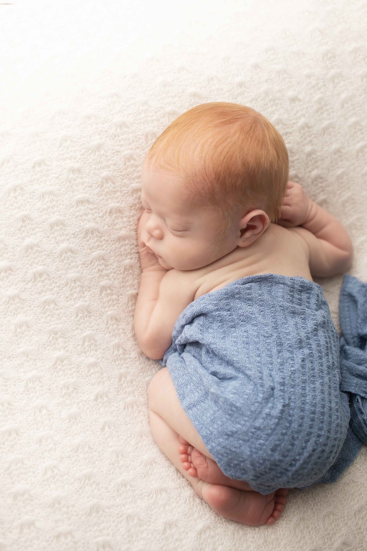 Newborn baby boy with fiery red hair resting on a simple backdrop, his tiny fingers gently curled