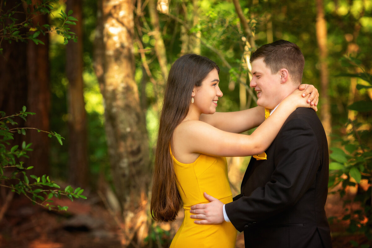 Prom couple in high school.  The girl and boy are facing each other.  She has her arms around his neck and he is holding her waist.  He is wearing a black suit and she is wearing a yellow dress.  She has long brown hair.  They are smiling at each other.
