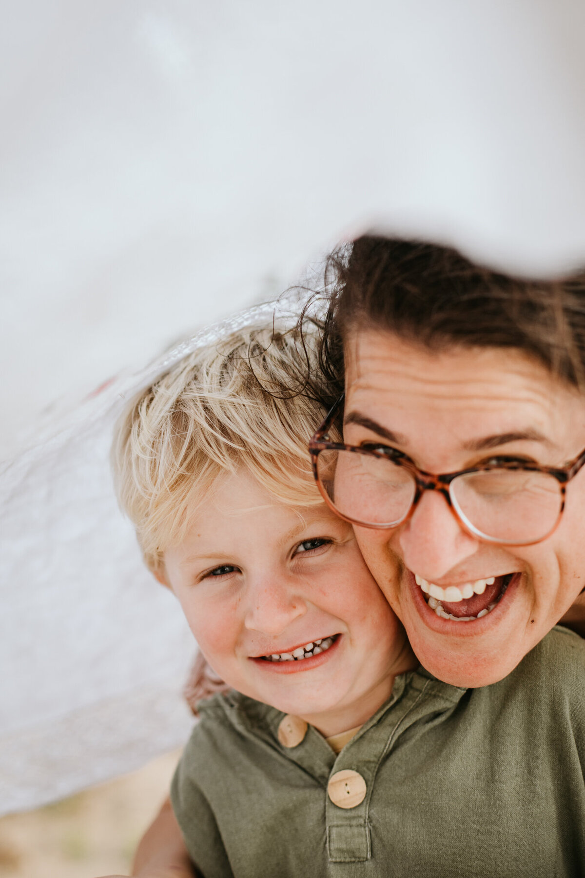 Mother and son peek out from under a linen sheet, facing the camera with playful expressions.