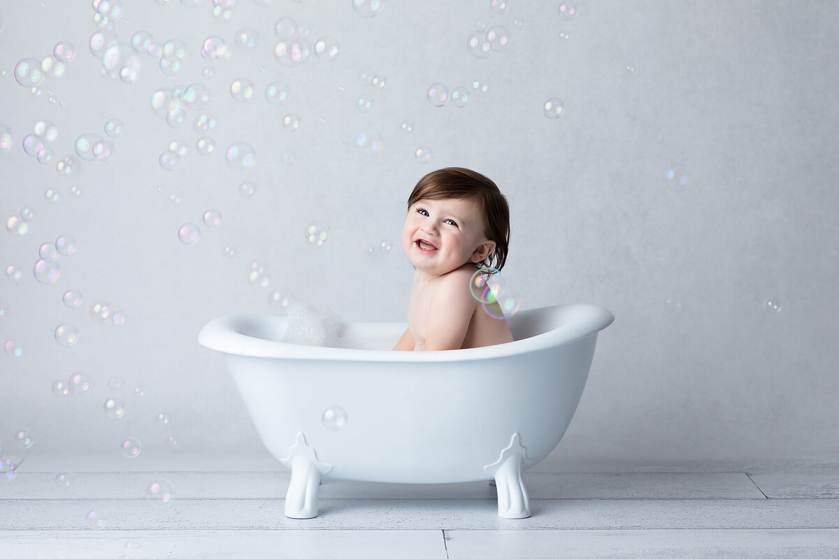 A joyful baby splashes in a white clawfoot tub surrounded by floating bubbles, captured during a whimsical Dallas milestone photography session.
