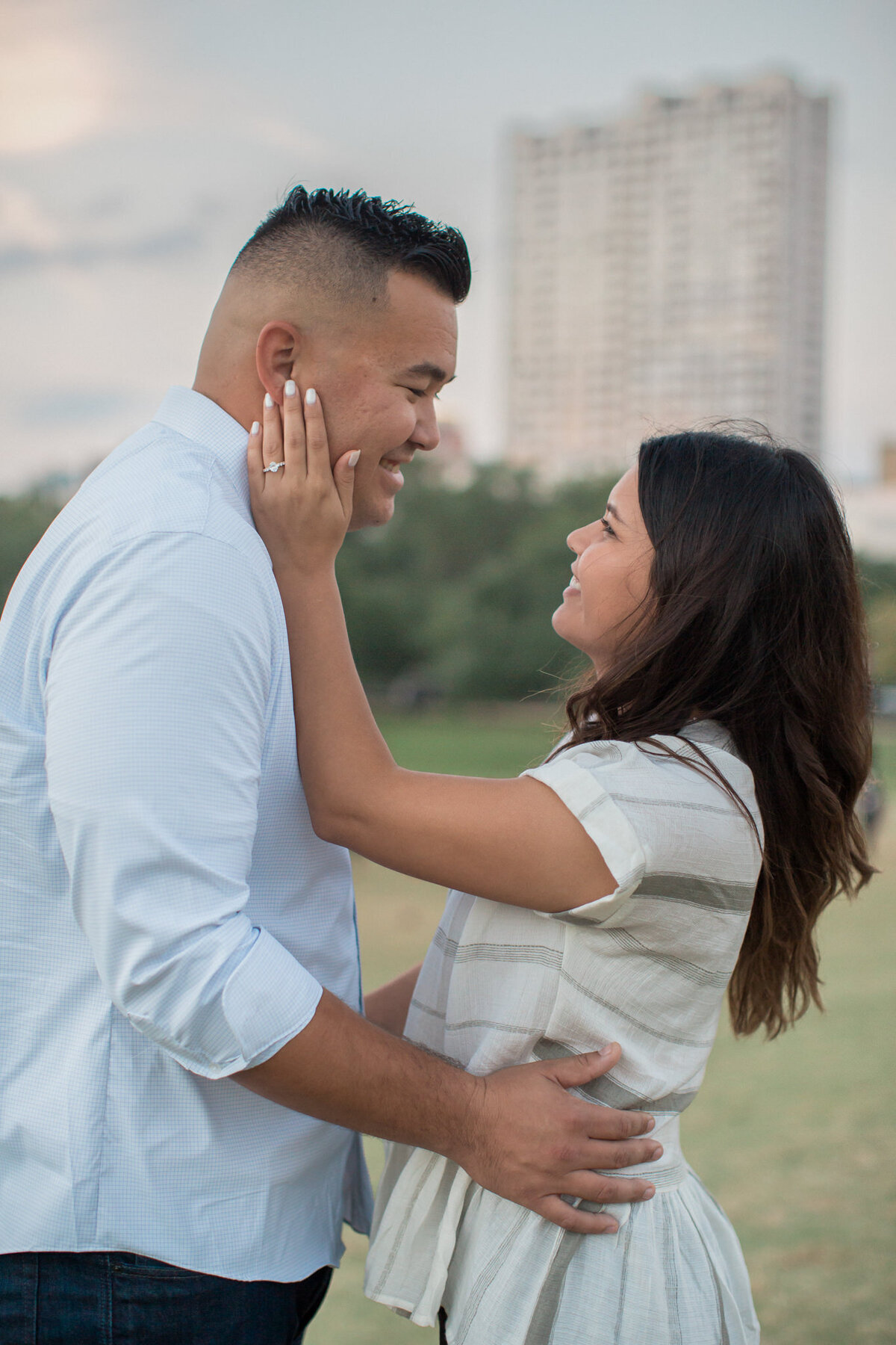 A latino woman places her hand on her fiancé's face, displaying her ring, with the Houston skyline in the background