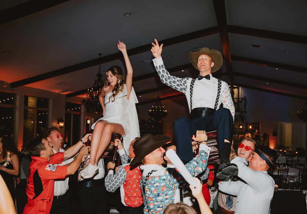 Bride and groom lifted on chairs during lively wedding reception celebration with guests dancing and cheering.