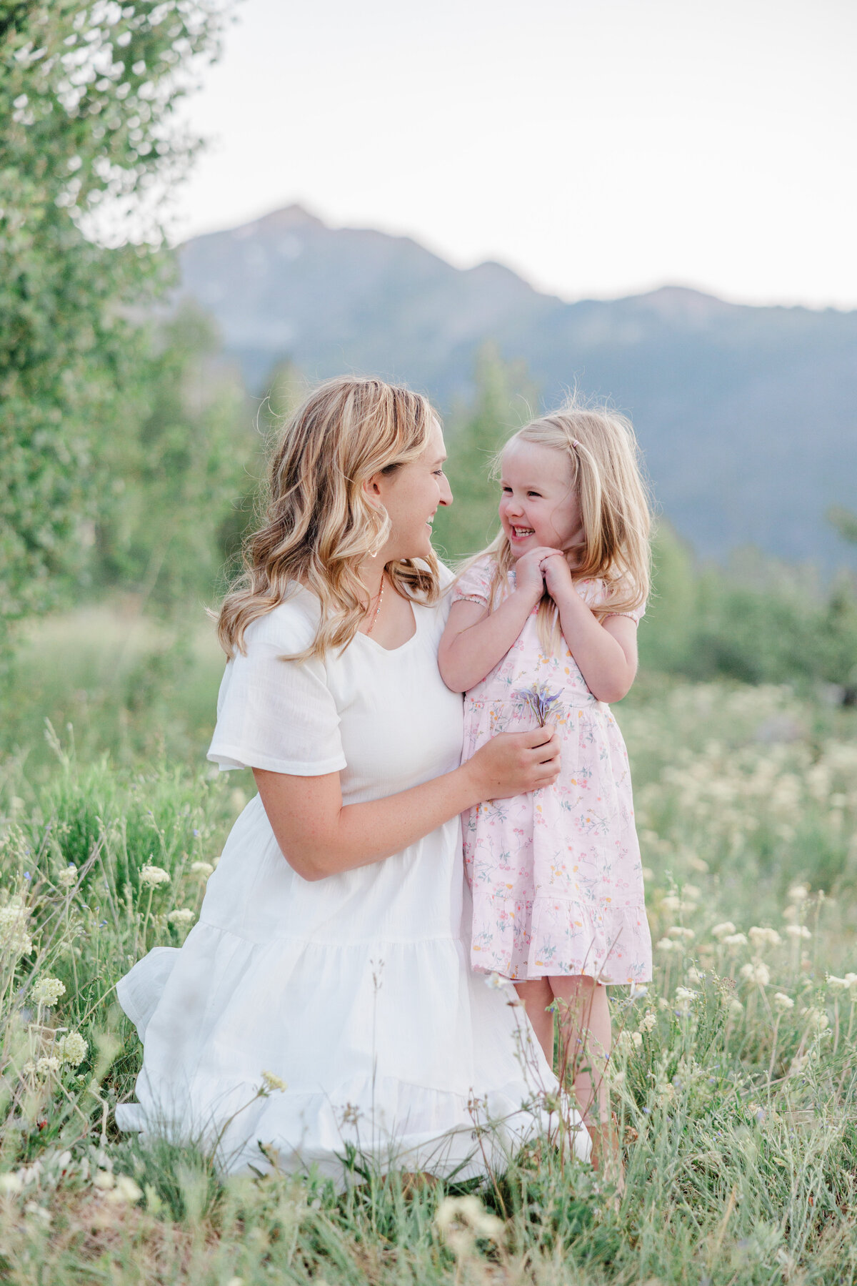 A happy mom laughs with her toddler daughter in a pink dress in a mountain meadow