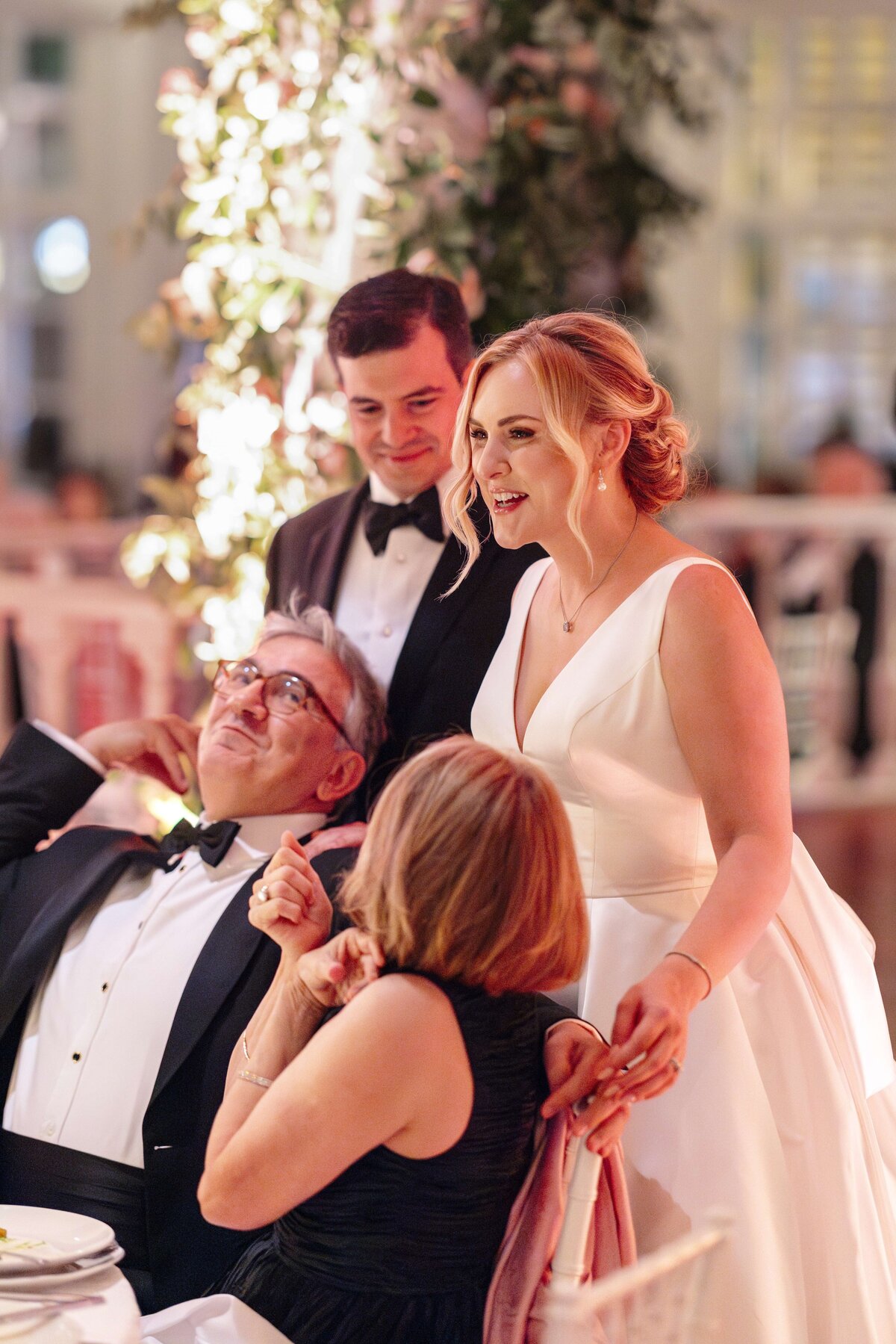 A smiling bride in a white gown stands beside seated guests at a wedding reception. A man in a tuxedo stands nearby. The scene is warmly lit with floral decorations in the background.
