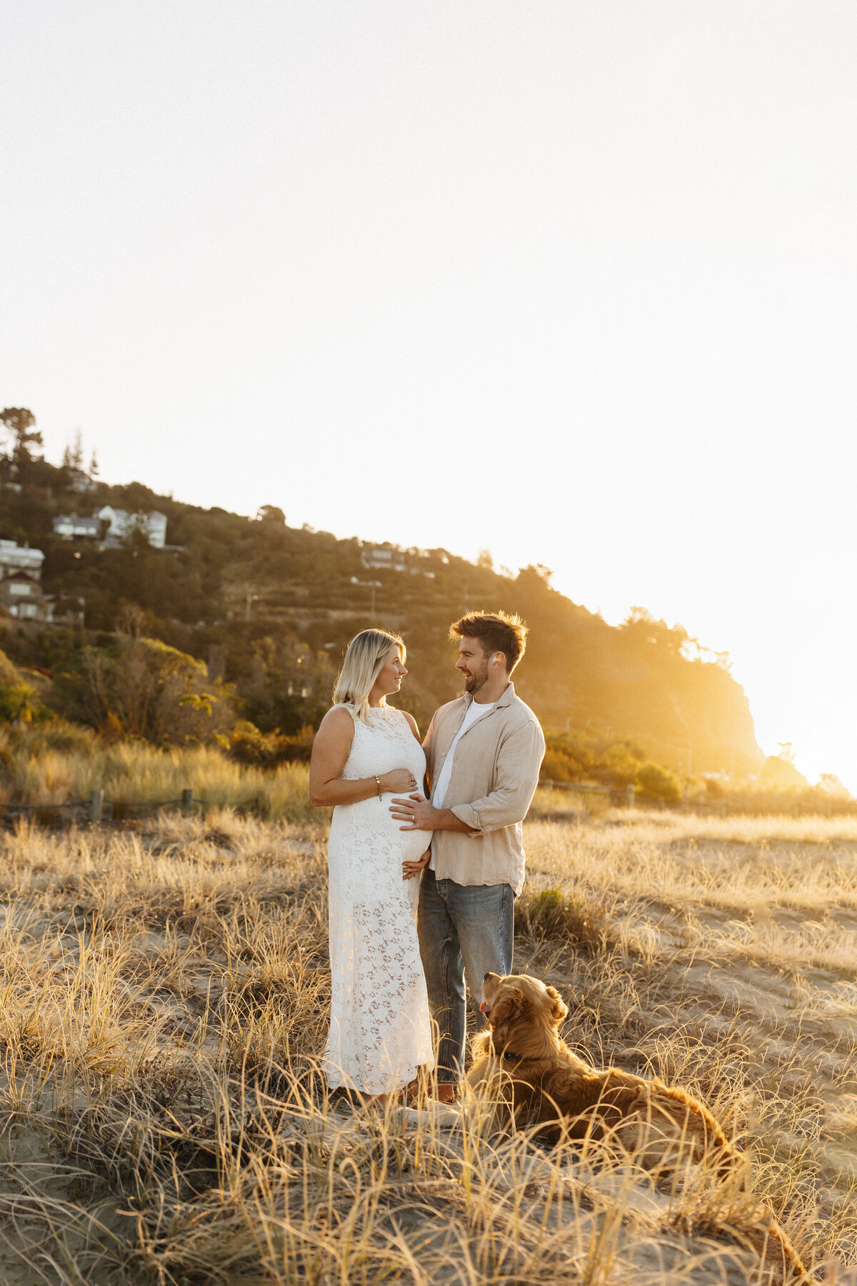 A mother to be and her partner hold her pregnant belly while posing with their family dog during their family maternity photoshoot in christchurch