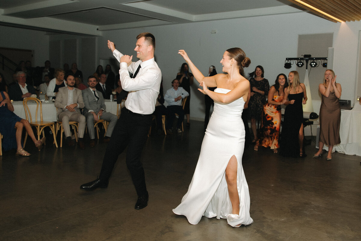 A bride in a white strapless, fitted wedding dress performs the thriller dance next to her groom. He wears a formal white dress shirt, black dress slacks and a black tie. He also performs the thriller dance in sync with his bride with both of their arms raised up to their right. Wedding guests stand and sit behind the couple watching them. 