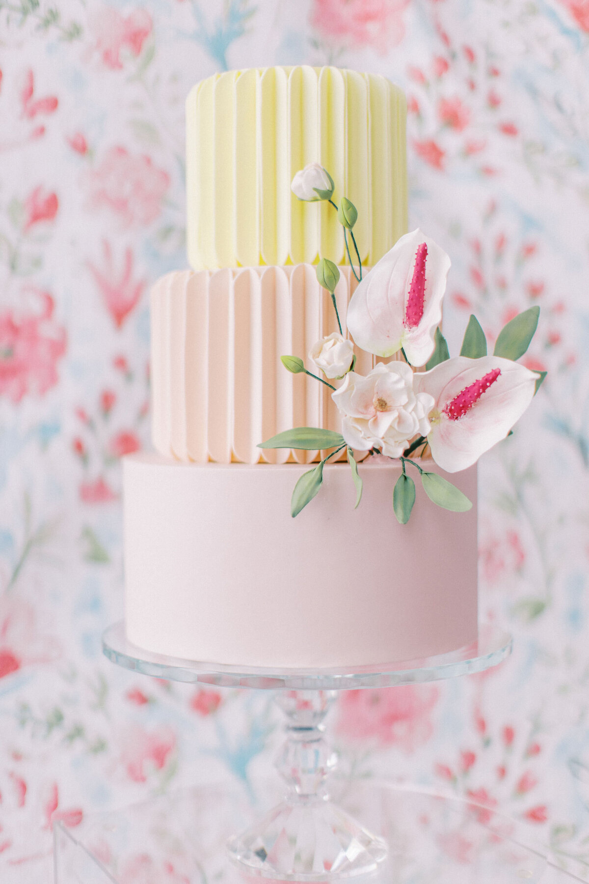 A three-tiered cake, decorated with pastel pleats and white-flowered accents, stands on a glass stand against a floral backdrop—a picture-perfect choice for weddings in Banff, Alberta.