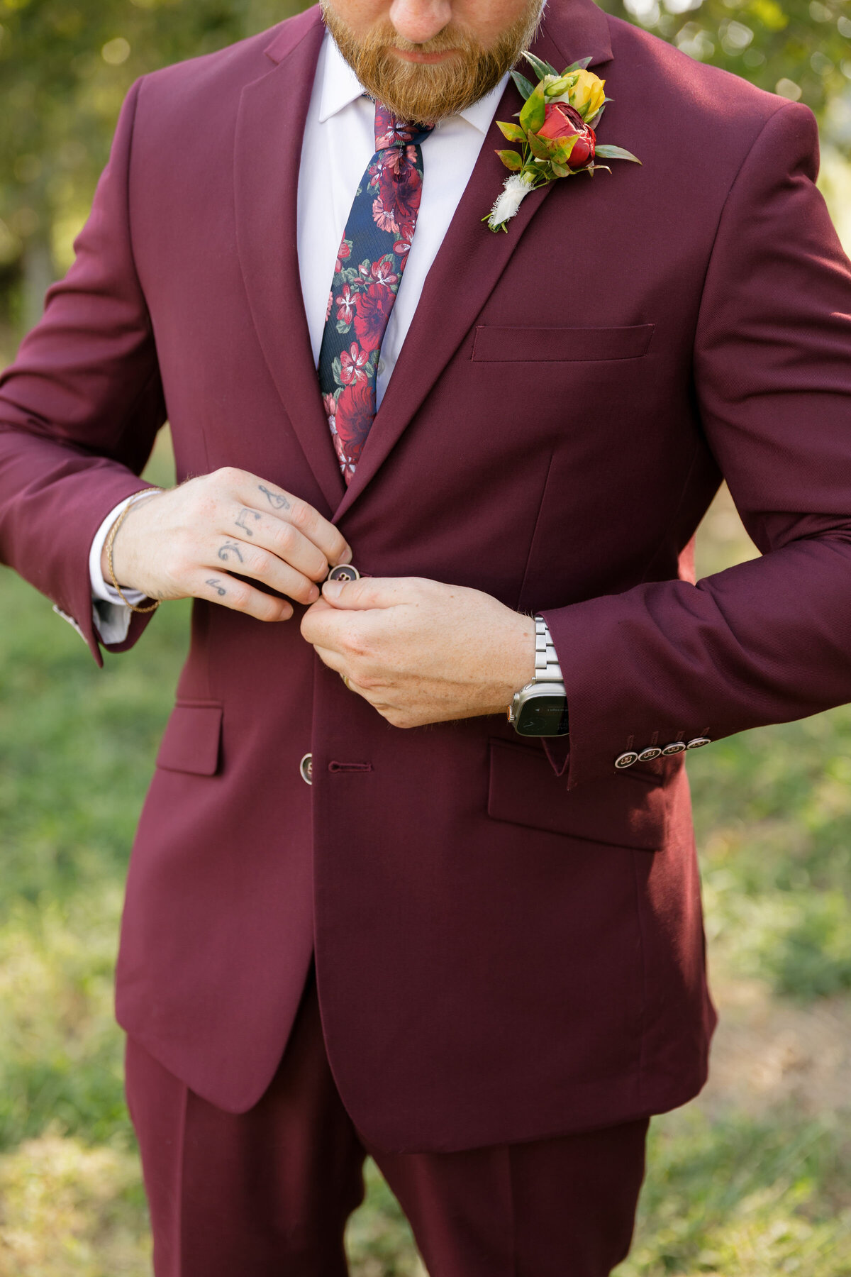 The groom buttons his burgundy suit jacket while standing outdoors before the wedding ceremony, showcasing his floral tie, wristwatch, and tattooed fingers. Soft natural light highlights the refined men’s wedding fashion and the deep tones of his suit, creating a polished groom portrait focused on attire and styling.