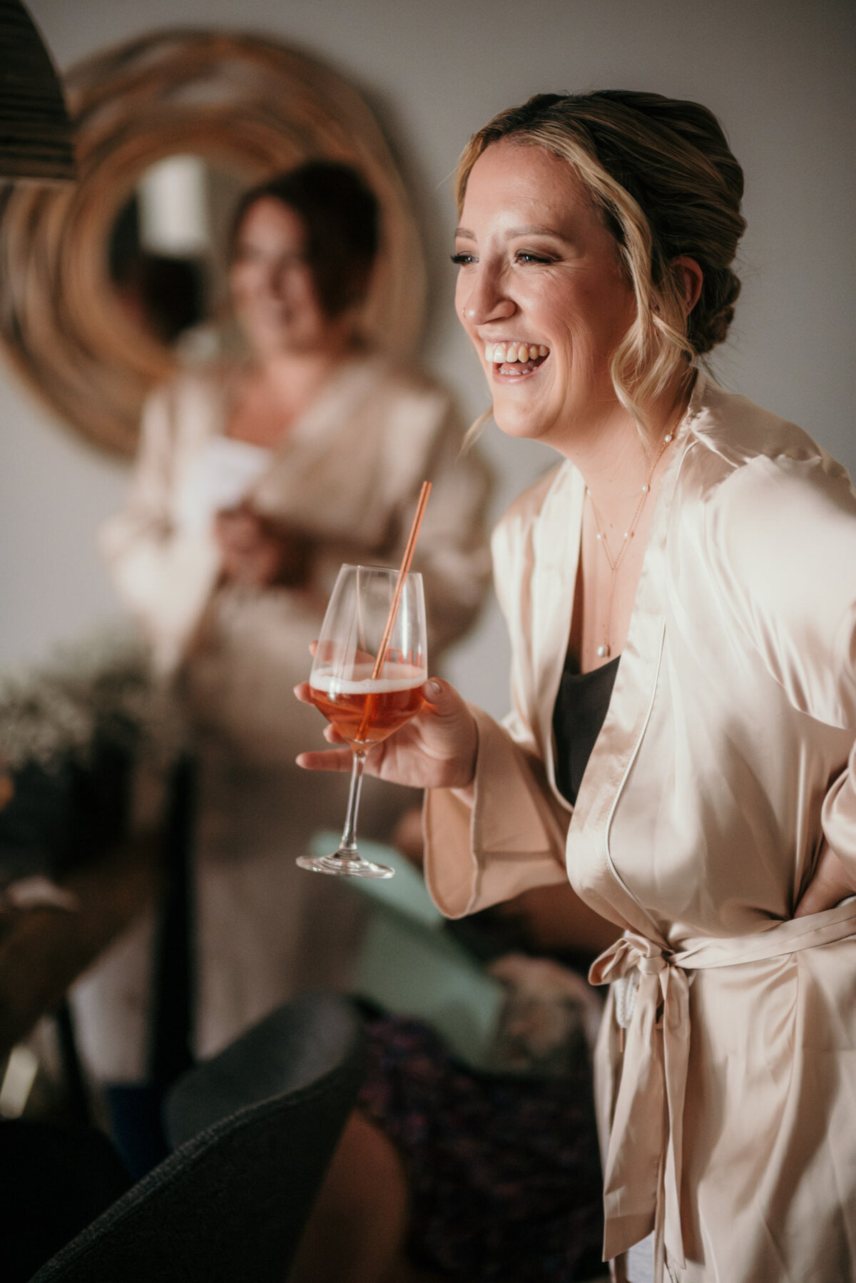 Eine der Brautjungfern in einem Satin-Bademantel lacht fröhlich mit einem Glas Sekt in der Hand.