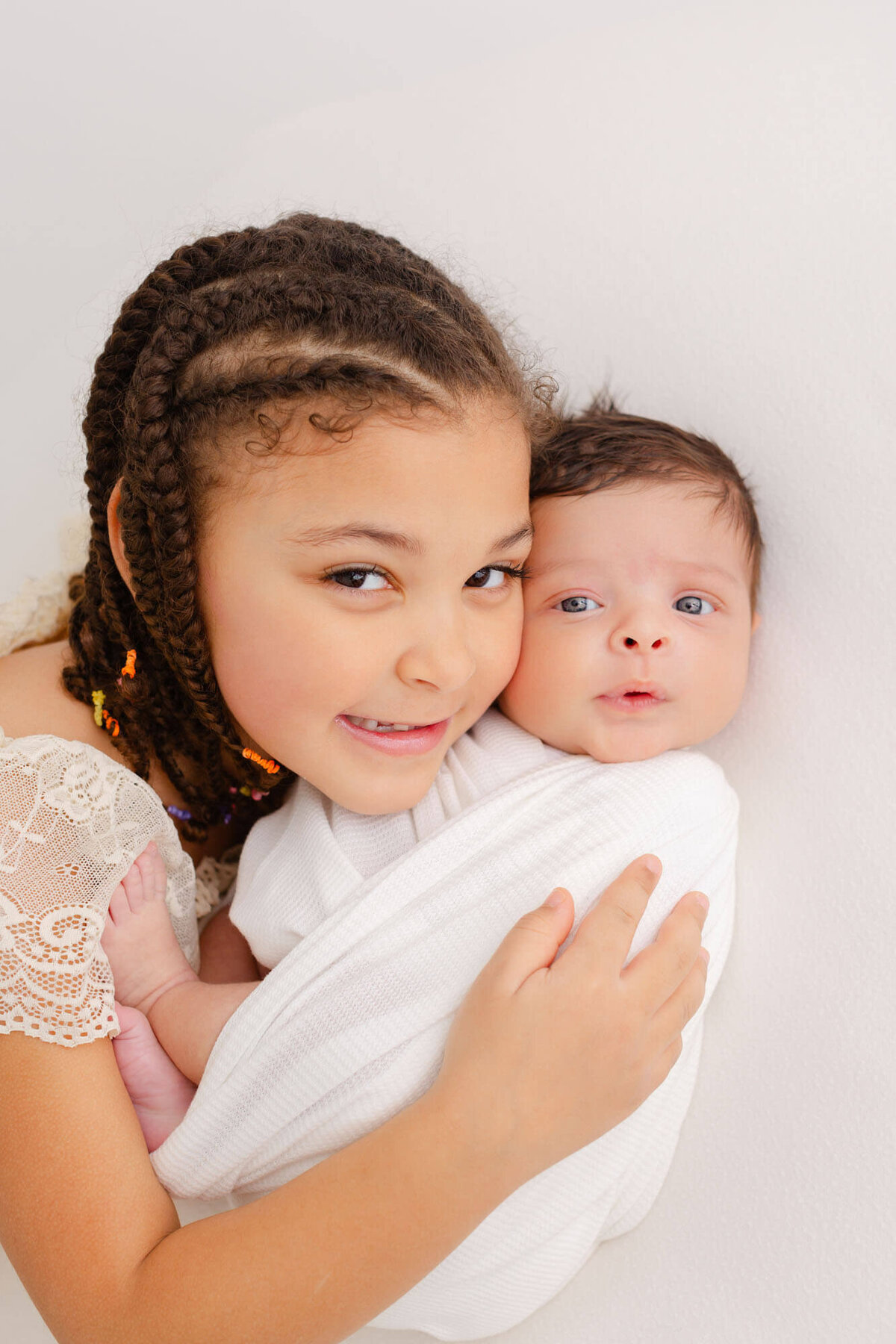 Big sister with hair in braids is cheek to cheek with her newborn baby brother. They both have their eyes open and are looking at the camera.