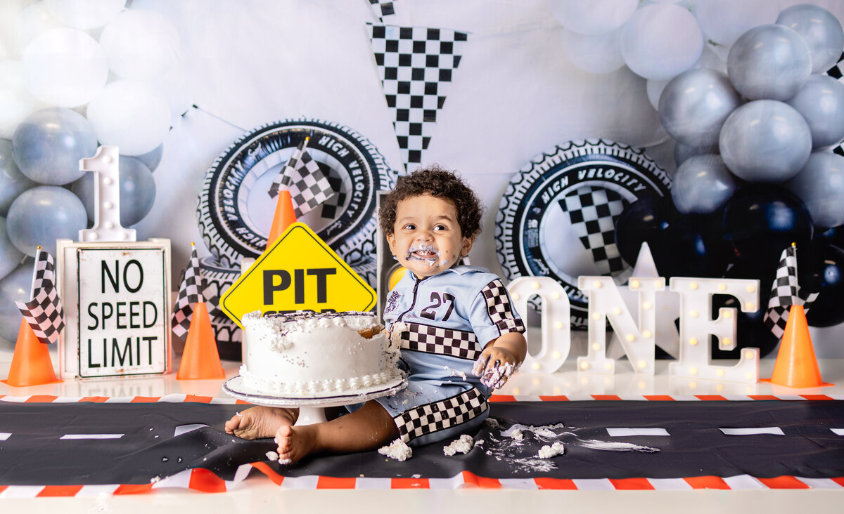 Race car themed cake smash with baby boy covered in frosting, sitting beside a smashed cake and checkered flags in Waldwick NJ"