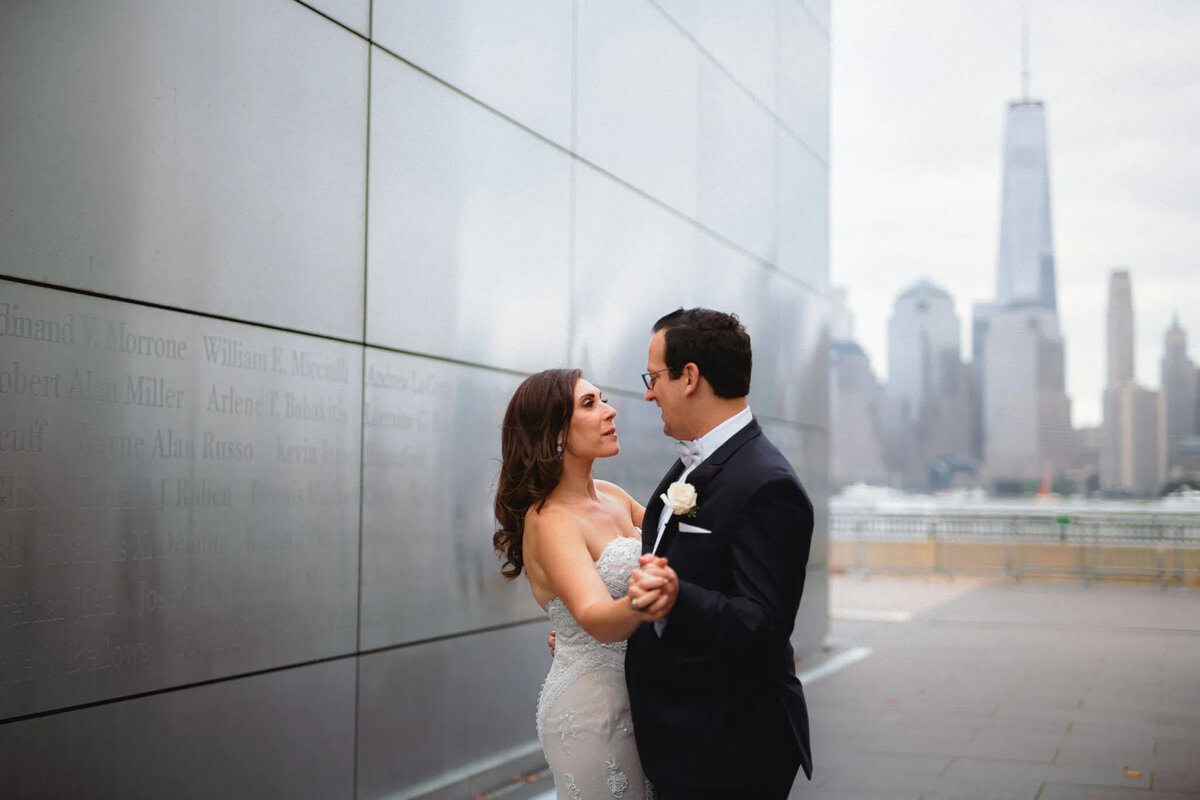 A bride and groom dance outdoors near a reflective wall as the New York City skyline, including One World Trade Center, shines in the background on a cloudy day, beautifully captured by an experienced NJ wedding photographer.