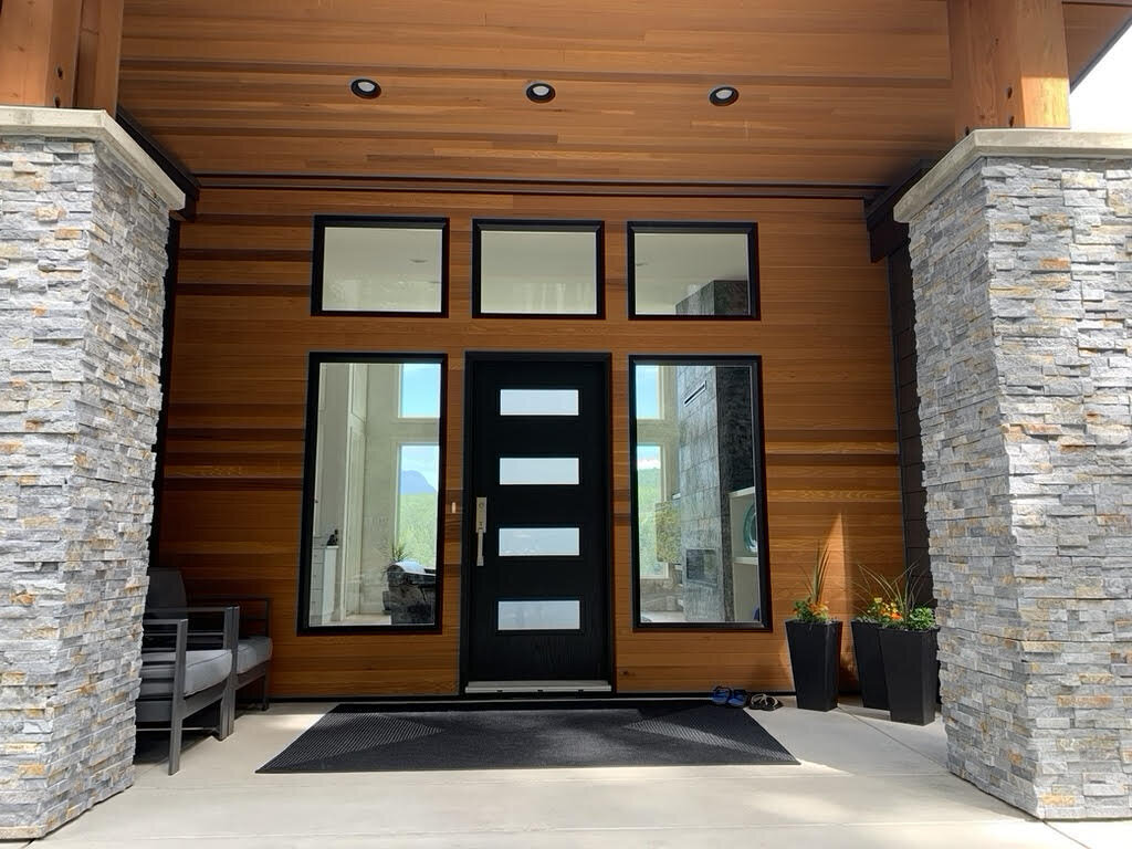 Modern home build entrance with plenty of windows, steel door, cedar soffits, and stone wrapped pillars.