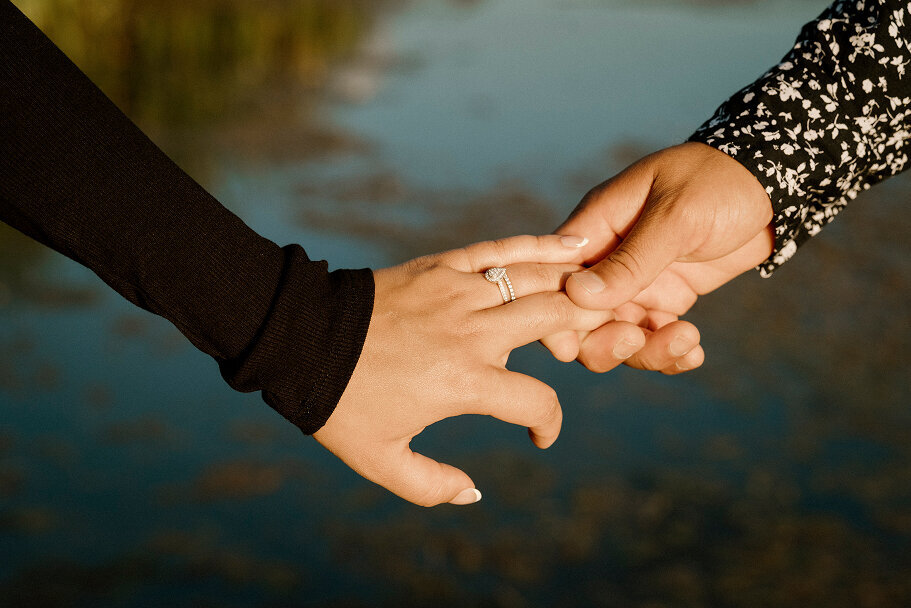 Close up shot of a man holding his fiancés hand. She is wearing her sparkly engagement ring. Behind them is a lake.