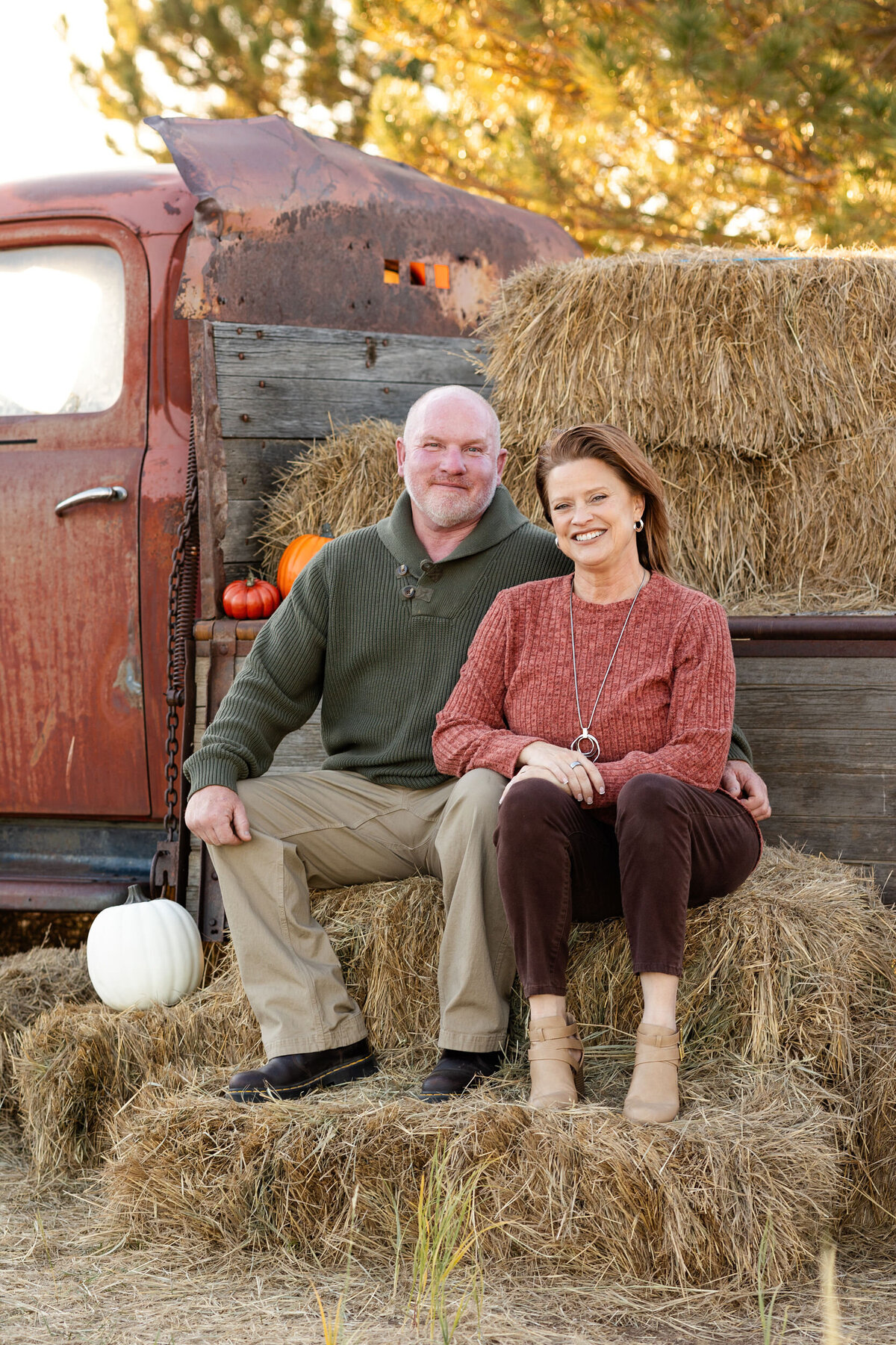 Husband and wife sit on a bale of hay in front of a vintage red farm truck and smile at the camera.