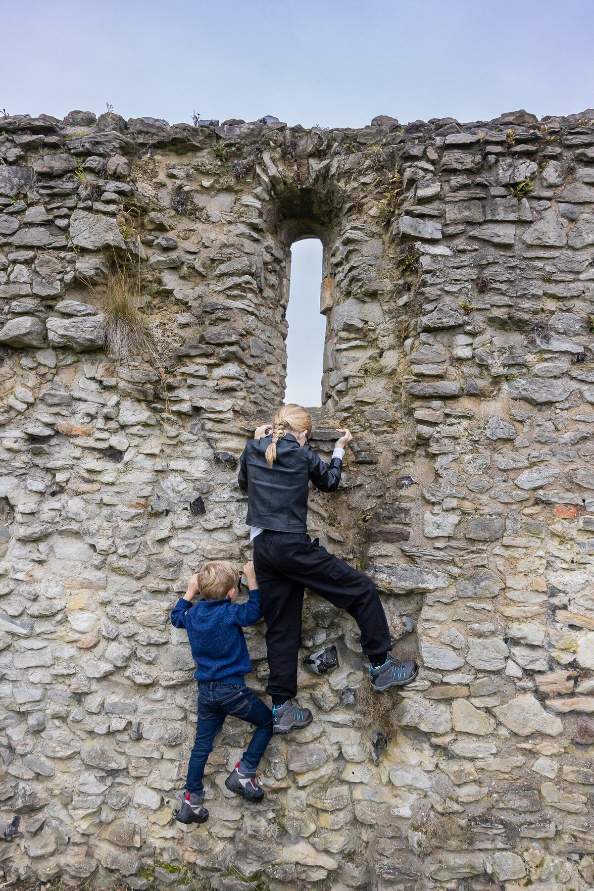 Two children climb an old stone wall, one reaching for a narrow vertical window, against a cloudy sky.