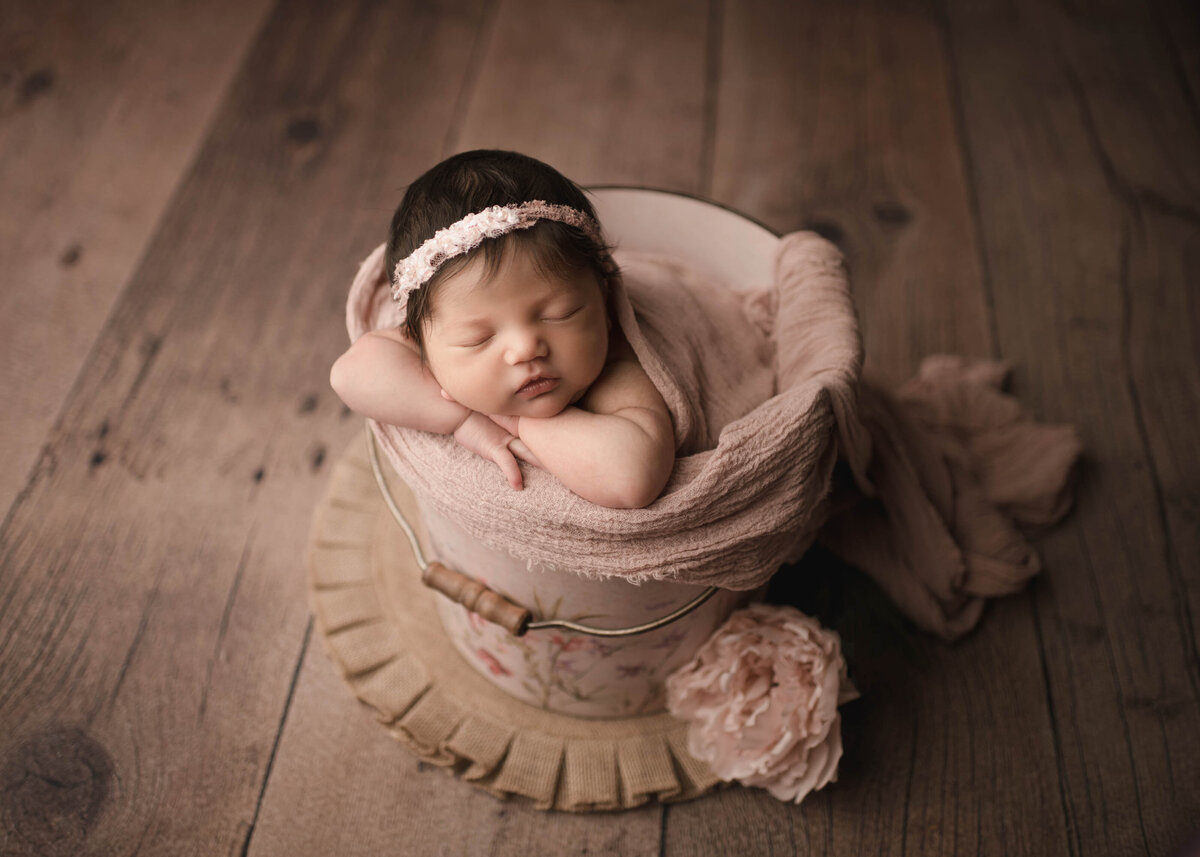 Baby girl is posed in a bucket for her newborn photoshoot. Her arms are folded on the edge of the bucket and her head is resting atop of her arms. She is sleeping peacefully. Captured by best Menifee Newborn Photographer Bonny Lynn Photography