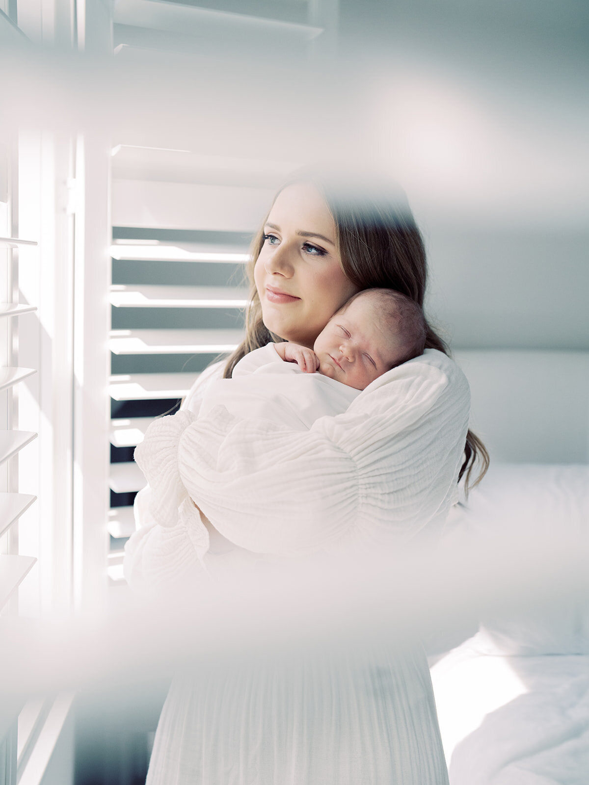 Mother with brown hair in white Nothing Fits But dress holds newborn baby on her chest as she gazes out the window by her blinds during her Bethesda newborn session.