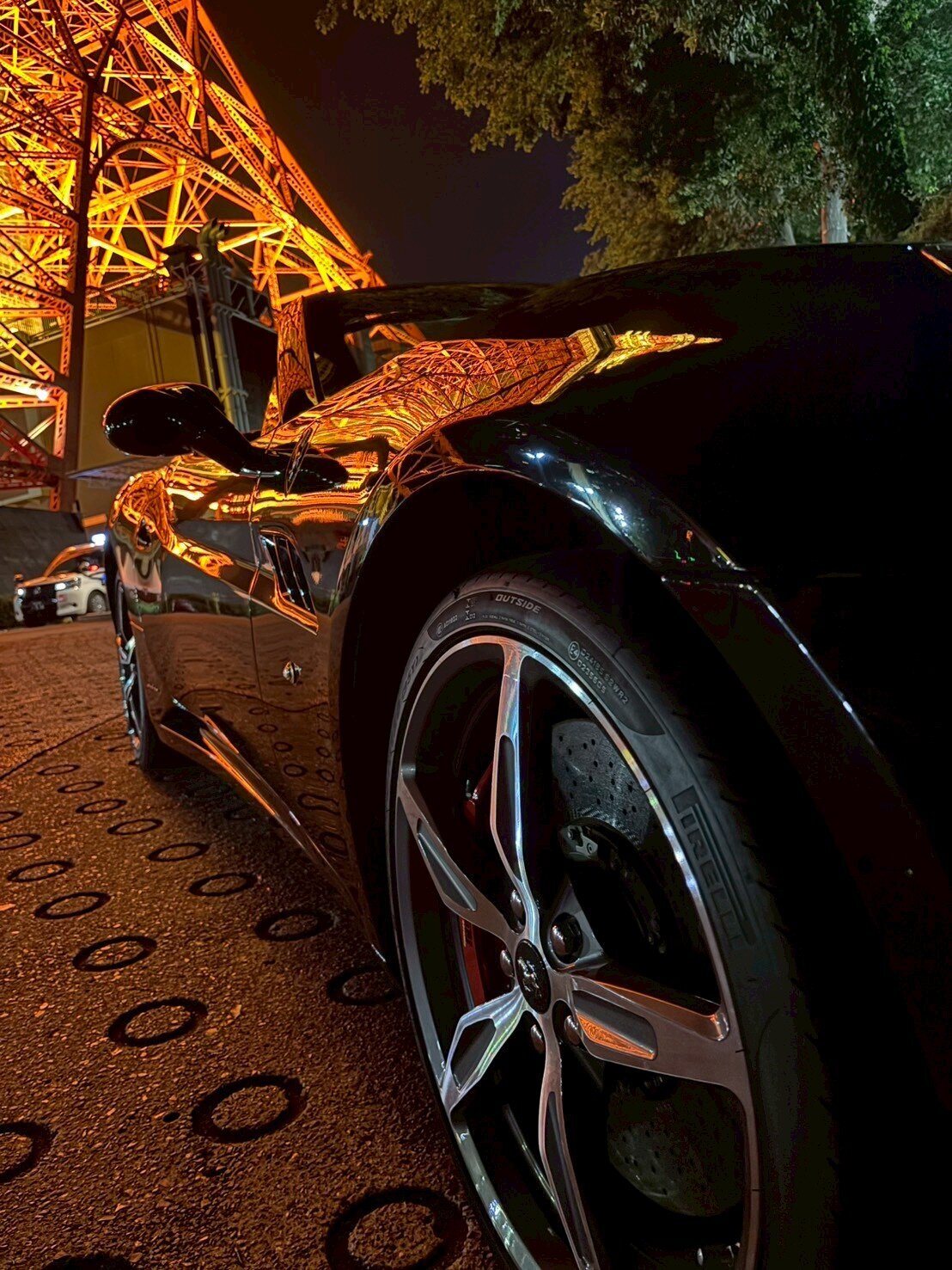 Close-up shot of a black Ferrari parked at night beneath Tokyo Tower, with the tower’s golden lights reflecting off the car’s polished body and chrome wheels.