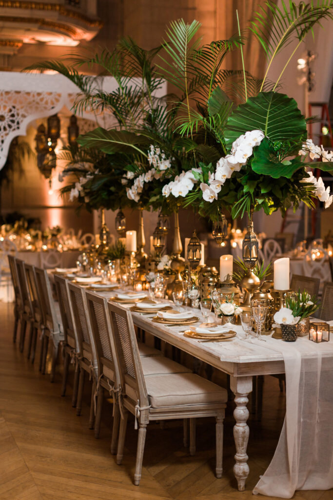 This stunning photo was taken at the Mellon Auditorium in Washington D.C. It showcases one of the best luxury DC venues with European flair captured beautifully by photographer Sarah Bradshaw. The image features a lavishly set dining table ready for a formal event, adorned with lush green foliage, white orchids, and elegant gold accents. The sophisticated ambiance is further enhanced by candles and decorative lanterns, while neatly arranged chairs and soft lighting provide a warm and inviting atmosphere.