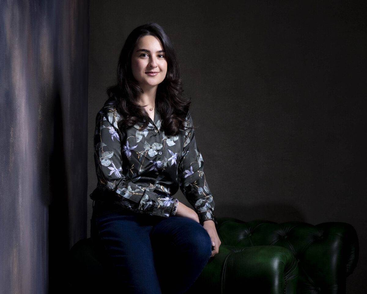 Environmental portrait of a woman seated on a green leather sofa, wearing a dark floral blouse, with soft side lighting and a dramatic backdrop, photographed by Rob Faber Photography.