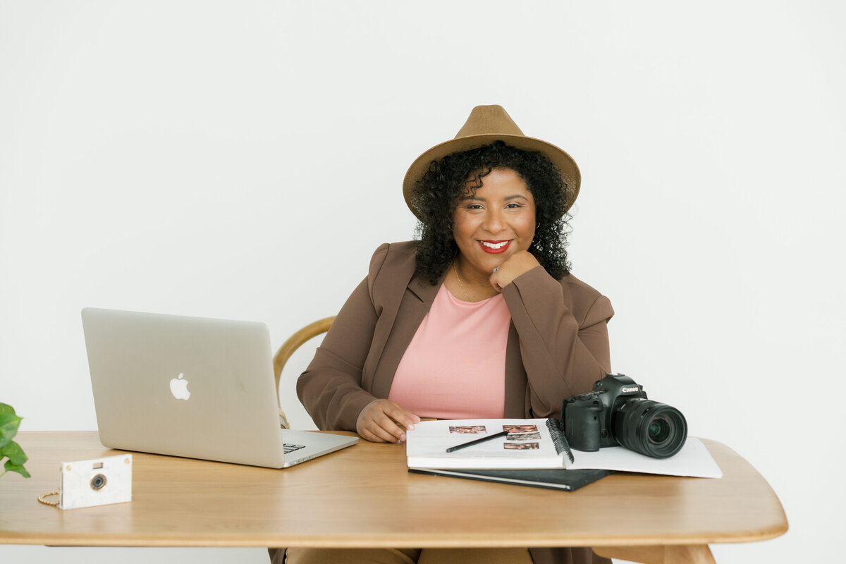 A professional yet approachable portrait of Mica McCook, an Austin-based food photographer, sitting at a desk with her camera, laptop, and portfolio. Mica’s warm smile and creative setup reflect her passion for photography and her engaging personality.