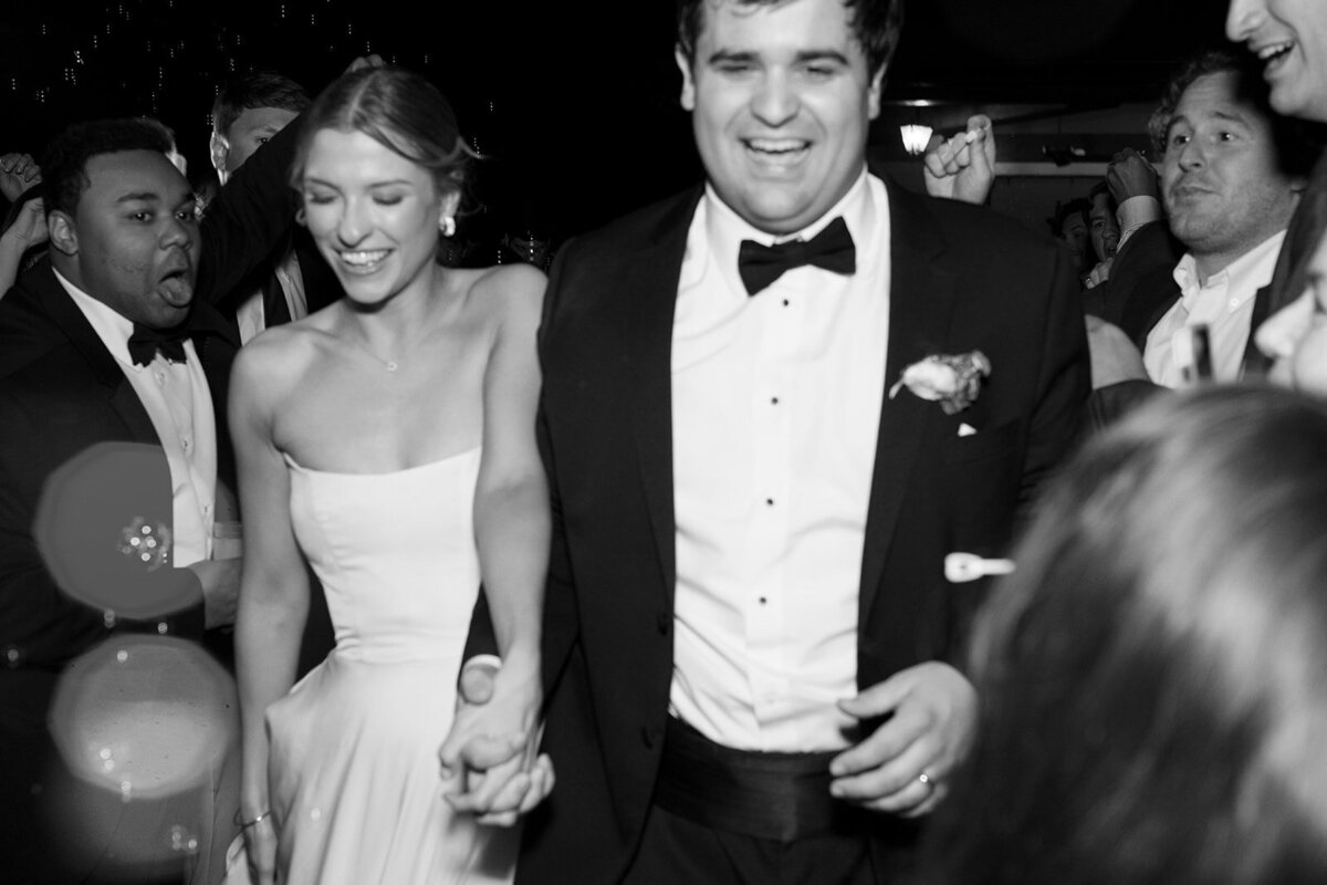 Black and white photo of Callie and Hayes walking hand-in-hand, smiling joyfully, during their Burritt on the Mountain wedding celebration.