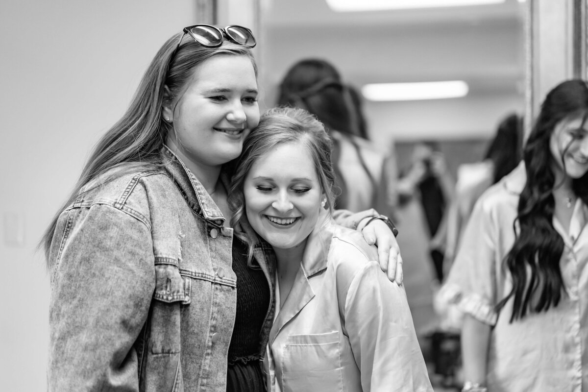 Two women smile as they pose together, one with her arm around the other. Both appear happy; one is wearing a denim jacket, the other light-colored pajamas. Blurred people are visible in the background. The photo is black and white.