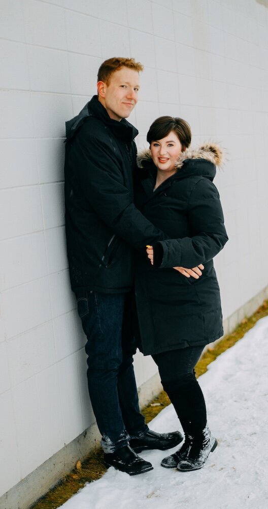 A boyfriend leans against a white wall while embracing his girlfriend