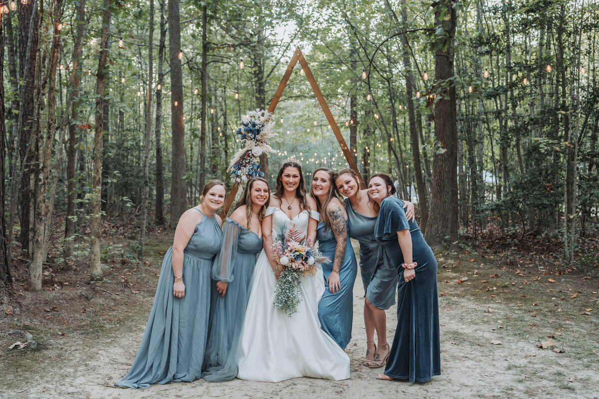A bride in a white wedding dress and her bridesmaids in blue dresses pose together in a forest setting under a wooden triangular arch adorned with flowers, with hanging string lights in the background. Shot by Quinn Photography LLC.