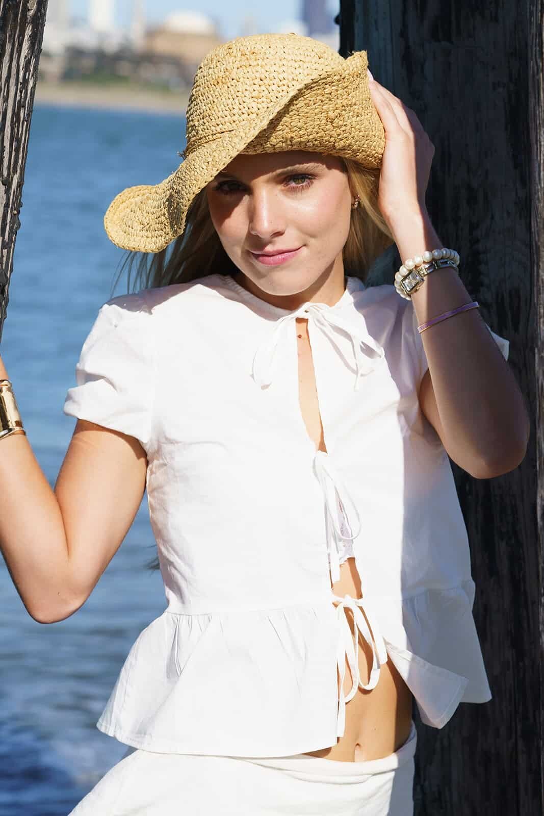 Young woman in a white top and a straw hat at the edge of the water smiling for a photo