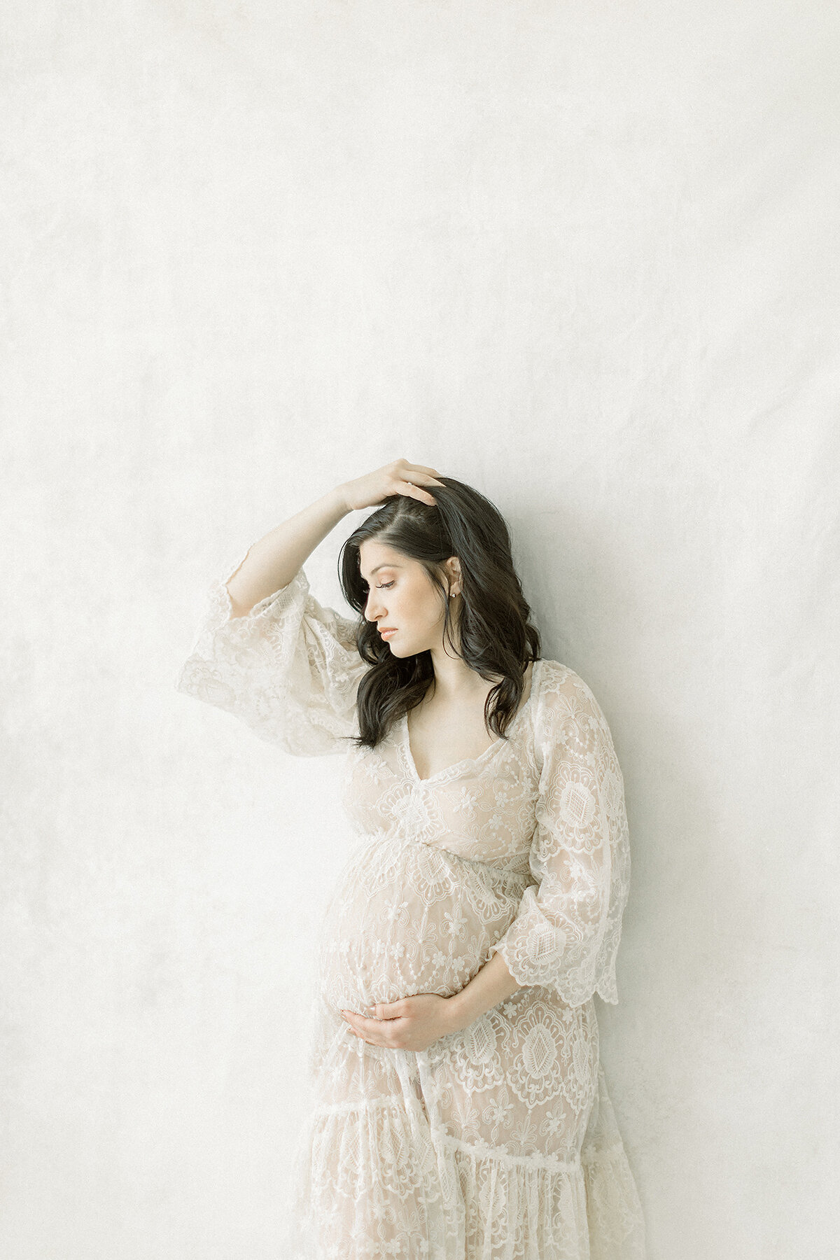 A maternity photo of a mother posing in a Dallas photography studio by the window as she is gently holding her hair with one hand and the other holding her belly.