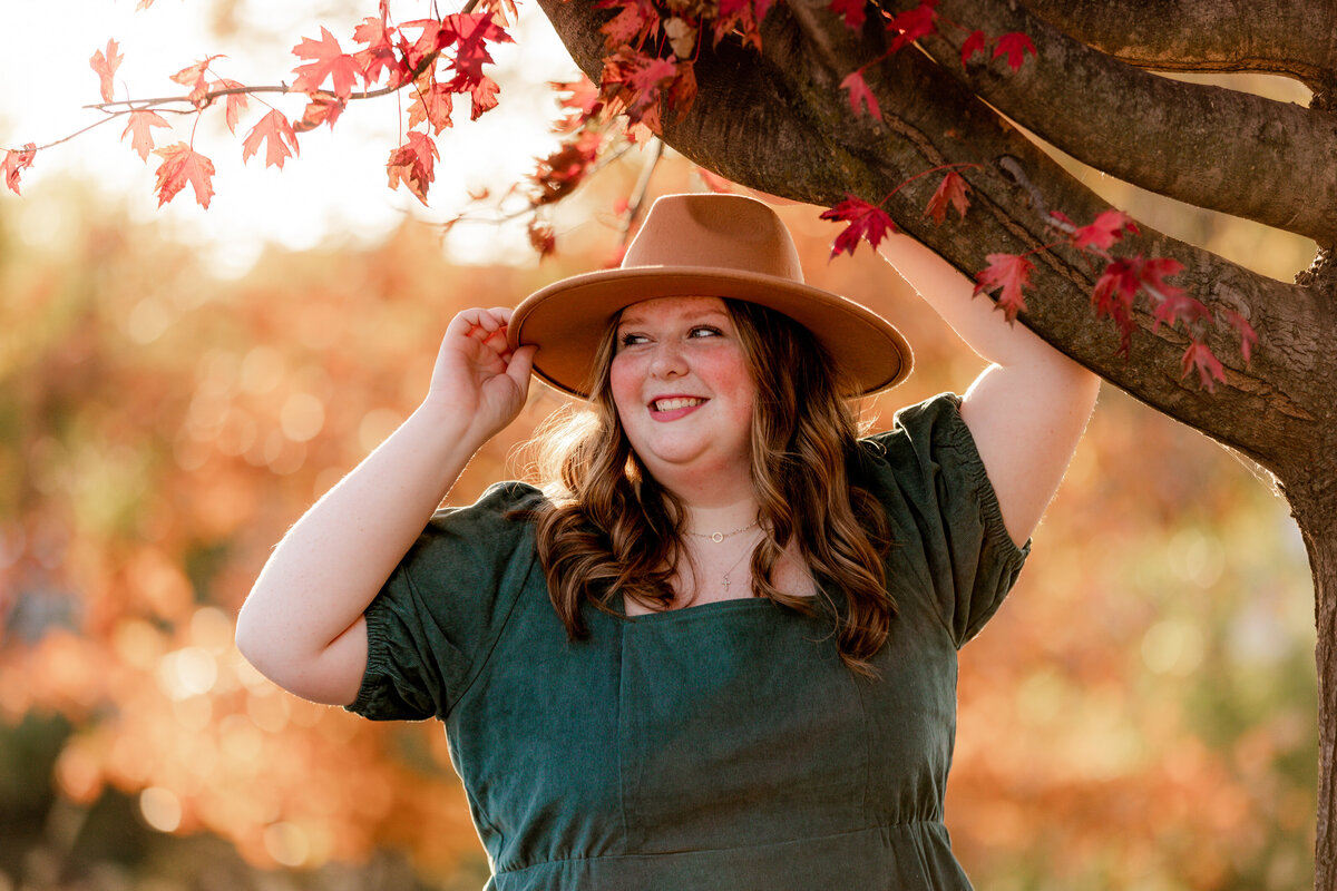 Calire holds one hand to her hat and her other hand up on a tree branch while she looks off into the distance.