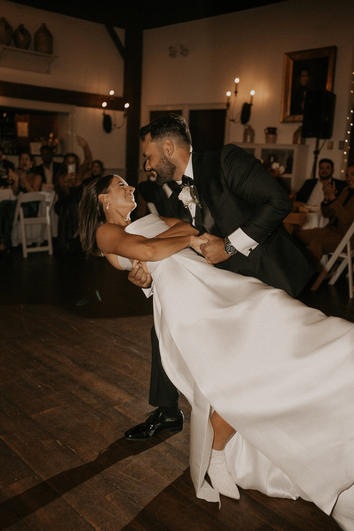 Bride and groom share a joyful dip on the dance floor during their wedding reception. The bride, in an elegant white gown, and the groom, in a black tuxedo, smile and laugh as they dance. The background shows cheering guests, capturing the lively and celebratory atmosphere of the evening.
