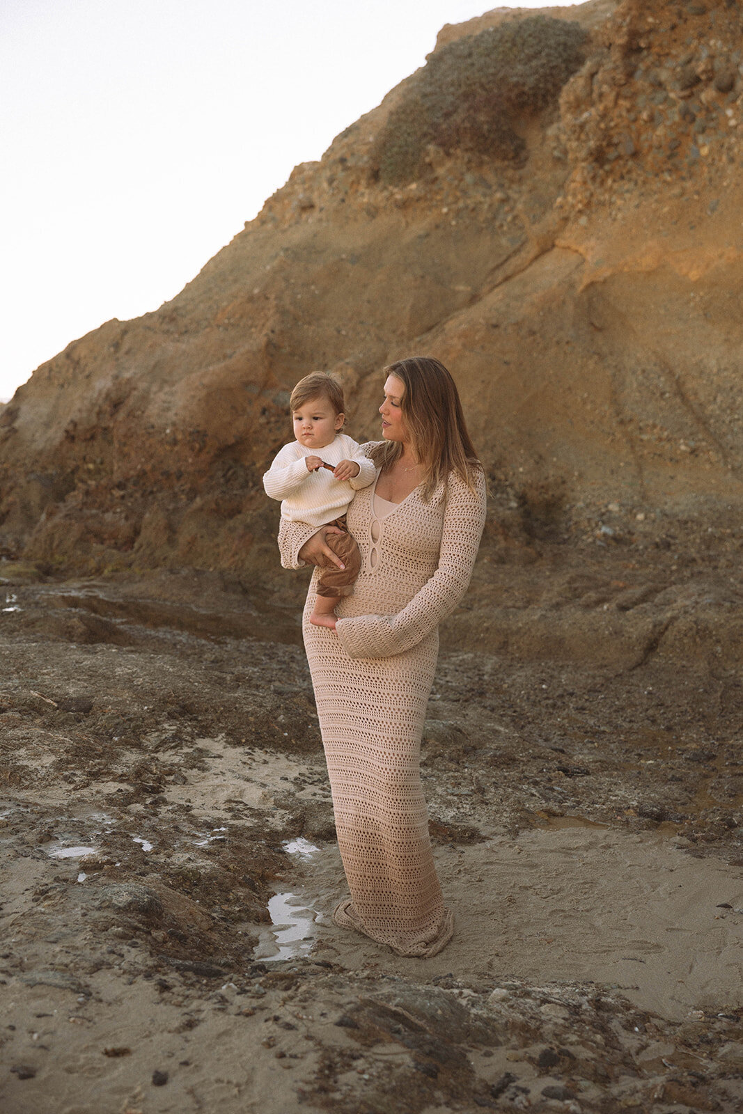 Mother in a neutral knit dress holding her child along the rocky shoreline at Goff Cove, part of an Orange County family photo session.
