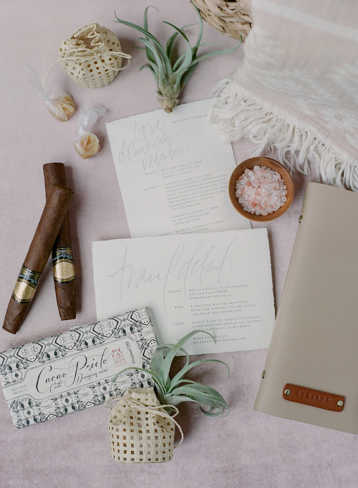 Flat lay image showcasing luxurious wedding details at a top tropical destination venue in the Caribbean, Dominican Republic. Items include two cigars, a bar of chocolate, air plants, packaged items, pink Himalayan salt, a SEYCHELLES labeled notebook, a fringed cloth, and elegant cursive stationery. Captured by photographer Laura Gordon.