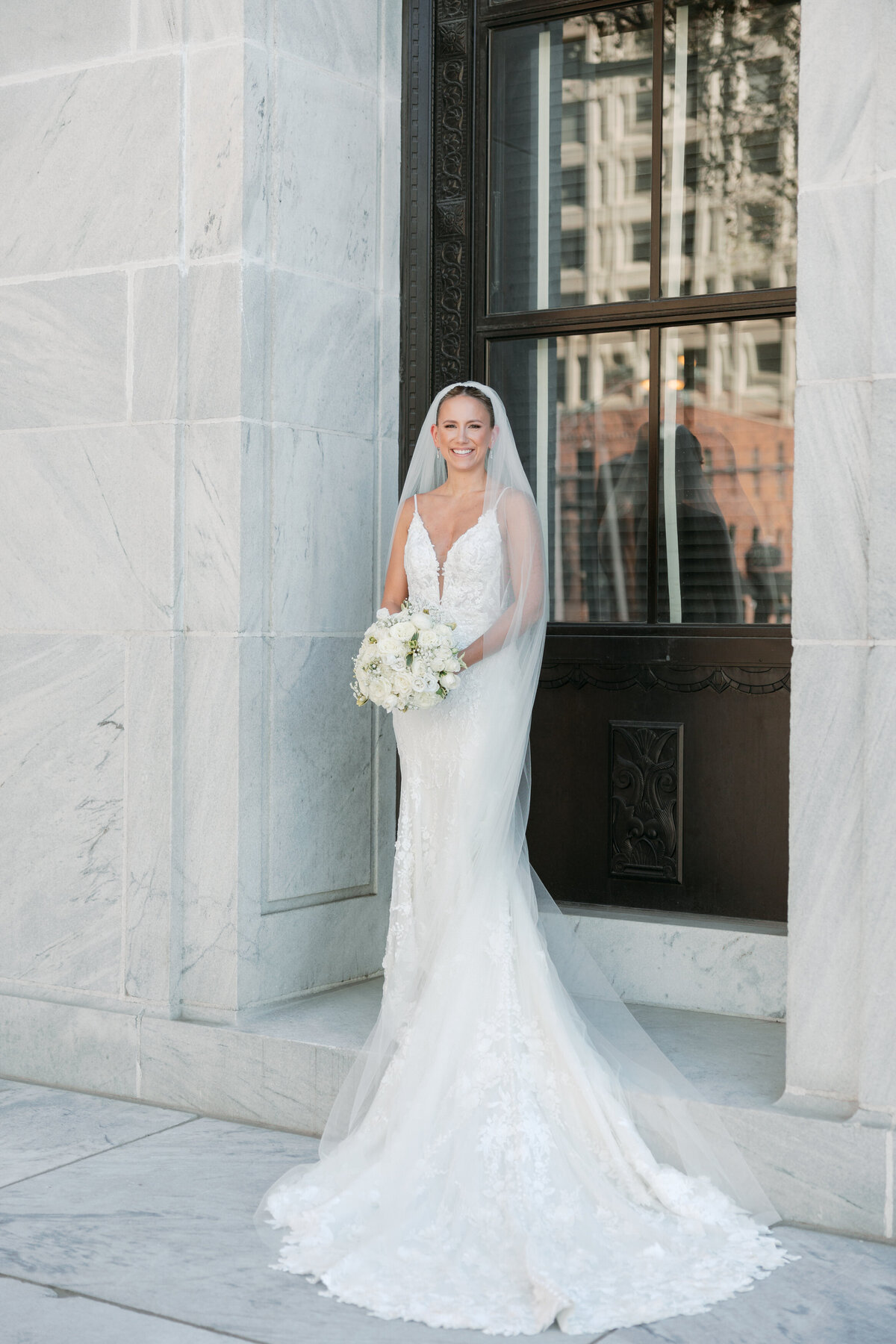 Bride standing alone in front of a window, holding bouquet and smiling, captured by a Philadelphia wedding photographer.