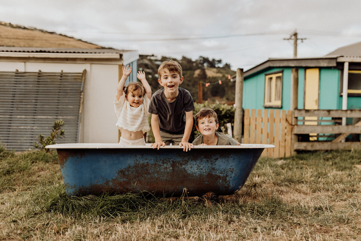 Three siblings laughing and jumping in an old bath tub during a family photoshoot with christchurch photographer lucy jane photography